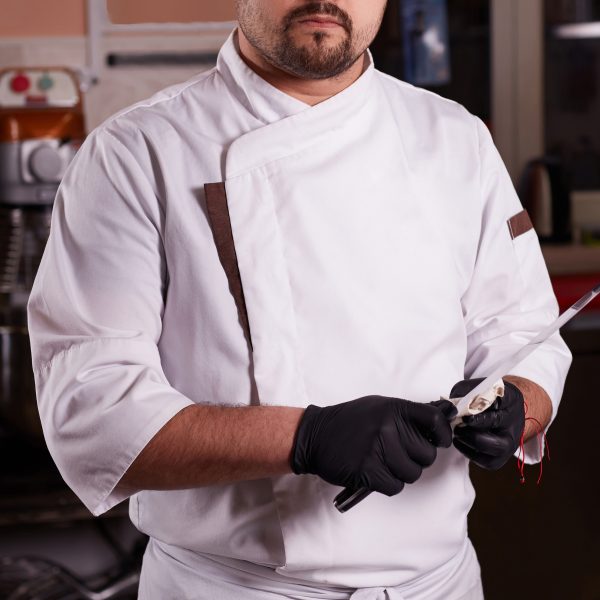 Portrait Of A Young Chef Cook In A Hotel Restaurant. Chef Is Cleaning A Knife.